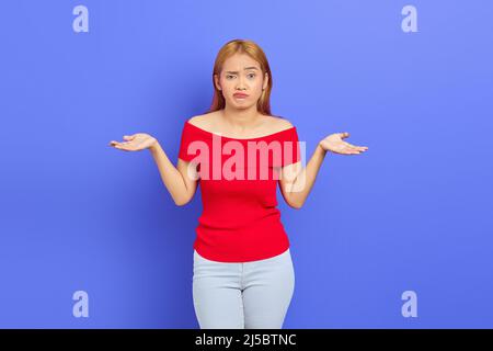 Portrait d'une jeune femme asiatique attirante et confuse avec des cheveux blonds courts en robe rouge debout et haussant les épaules isolées sur fond violet Banque D'Images