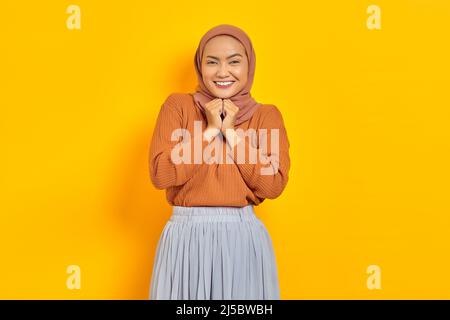 Belle femme asiatique souriante en chandail brun et hijab garde les mains sous le menton, regardant la caméra, se souvient agréable moment isolé sur bac jaune Banque D'Images