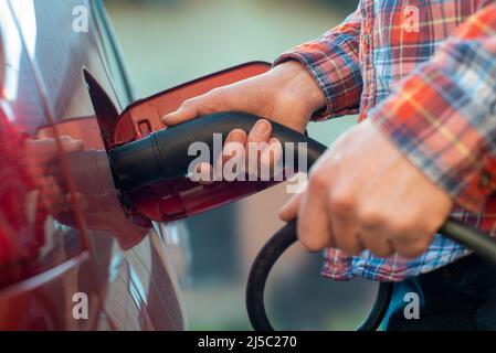 Le modèle mâle se tient près d'une voiture électrique et tient une fiche de charge Banque D'Images