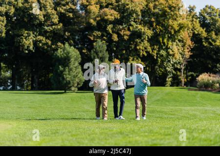 des amis multiethniques âgés souriant et marchant avec des clubs de golf sur le terrain vert Banque D'Images