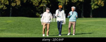 amis séniors interraciaux marchant avec des clubs de golf sur le terrain, bannière Banque D'Images