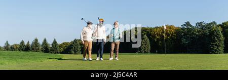 des amis interraciaux seniors marchant avec des clubs de golf sur le terrain vert, bannière Banque D'Images