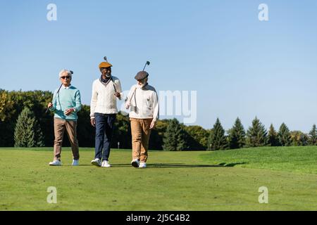 des amis interraciaux seniors marchent avec des clubs de golf sur le terrain vert Banque D'Images