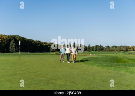 homme asiatique senior en lunettes de soleil marchant près d'amis interraciaux avec des clubs de golf Banque D'Images