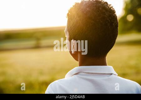 Garçon noir portant une chemise blanche regardant le coucher du soleil sur le terrain d'été Banque D'Images