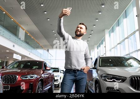 un homme d'âge moyen dans un concessionnaire de voitures tout-terrain prend un selfie entre les rangées de voitures Banque D'Images