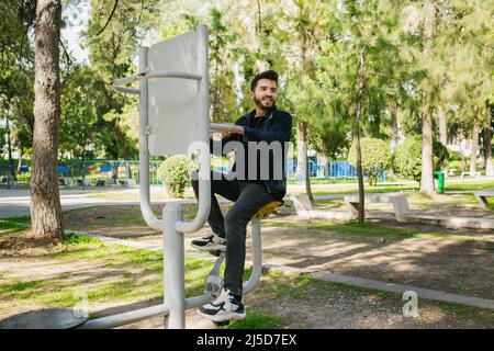 Exercice, jeune homme barbu à vélo dans des équipements sportifs publics, exercice court et vie saine, un parc vert et plaisir printanier Banque D'Images