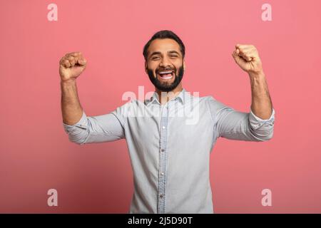 Un chanceux. Un gars indien excités célébrant la victoire et faisant la fête OUI avec des poings élevés sur fond rose. Bonhomme à barbe multiracial surjoyeusement en Jean décontracté gagnant de chemise Banque D'Images