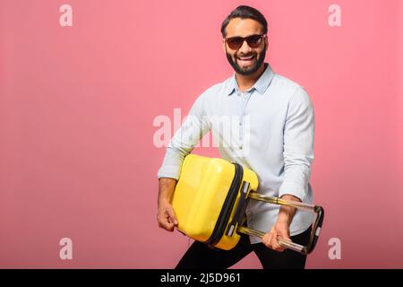 Un Indien joyeux et plein de joie dans des lunettes de soleil se tient avec une valise jaune, joue sur un sac comme un guitariste, se berner, un homme hispanique avec des bagages regardant un voyage, isolé sur le rose Banque D'Images