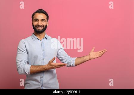 Un gars indien charmant pointe les paumes vers un espace de copie vide isolé sur fond rose. Joyeux homme multiracial barbu dans décontracté Jean chemise présentant la nouveauté, concept publicitaire Banque D'Images