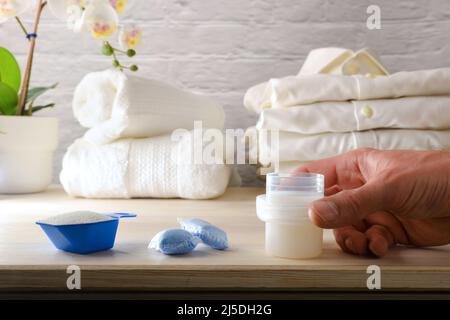 Main montrant différents produits chimiques de blanchisserie de ménage sur un banc en bois dans une buanderie à la maison avec des vêtements blancs fraîchement lavés à l'arrière-plan. Banque D'Images