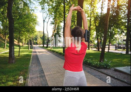 Vue du dos à une athlète musclée, sportswoman levant les bras, s'étirant et réchauffant son corps avant le jogging le matin le long de la ville pa Banque D'Images