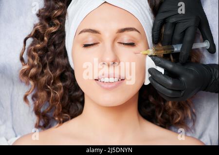 Gros plan des mains du médecin esthéticien dans des gants stériles faisant l'injection dans le visage de la jeune femme. Une femme porte un bandeau blanc tout en recevant un soin du visage au salon de beauté. Banque D'Images
