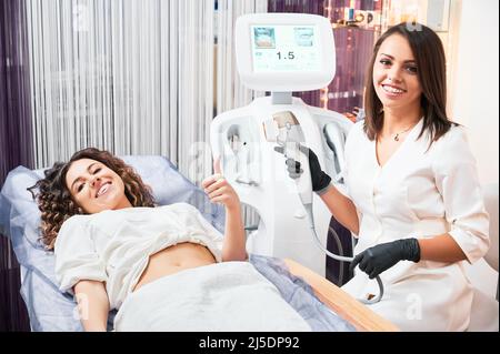 Patiente jeune femme couché montrant le pouce vers le haut et se préparant pour le diagnostic du corps avec l'équipement d'échographie. Concept de l'examen de santé de processus en clinique avec des équipements modernes. Banque D'Images