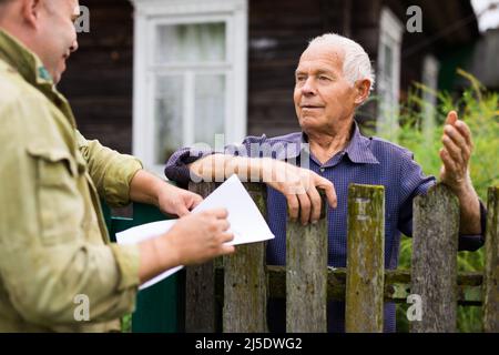 Homme senior communiquant avec le représentant de la compagnie d'assurance tout en se tenant à la clôture de sa maison de campagne Banque D'Images