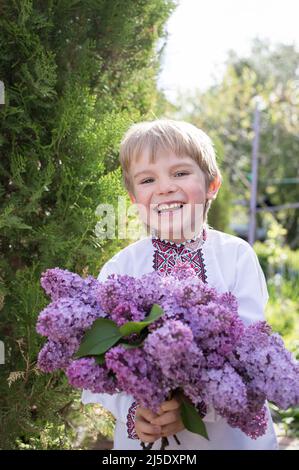 Un garçon ukrainien souriant de 4-5 ans dans une chemise brodée avec un grand bouquet de lilas. Fête des mères, bouquet de fleurs du fils. Fierté, liberté, moi Banque D'Images