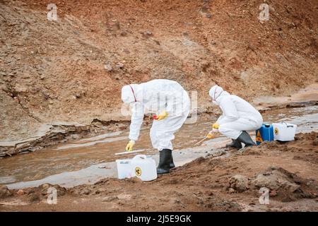 Deux spécialistes de l'environnement en vêtements de travail et masques de protection qui prennent des échantillons d'eau toxique tout en l'examinant sur une zone polluée Banque D'Images