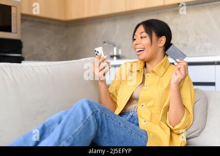 Photo d'une jeune femme excitée qui fait des achats en ligne tant attendus, heureuse femme tient un smartphone et une carte de crédit, magasiner en ligne, rêver avec les yeux fermés au sujet de nouvelles choses. Concept de consumérisme Banque D'Images