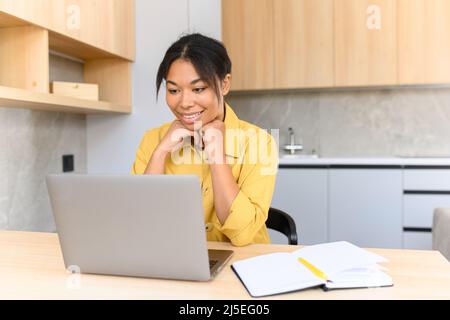 Une jeune fille multiraciale gaie utilise un ordinateur portable pour travailler à distance ou pour se divertir à la maison tout en étant assise dans la cuisine à la maison. Vue latérale une belle fille avec une coiffure afro regarde l'écran avec un sourire Banque D'Images