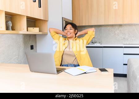 Sereine femme afro-américaine utilisant un ordinateur portable pour travailler ou étudier à distance, assise à la table et au repos, tenant les mains derrière la tête, passe un week-end paresseux à la maison, rêvant de jour Banque D'Images