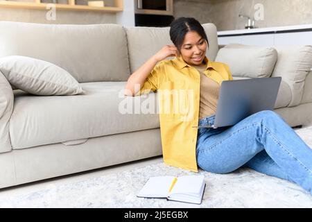 Charmante et calme, cette femme afro-américaine est assise sur le sol dans un salon confortable avec un ordinateur portable. Elle aime passer un week-end paresseux dans une atmosphère détendue, travailler à distance à la maison, discuter ou étudier en ligne Banque D'Images