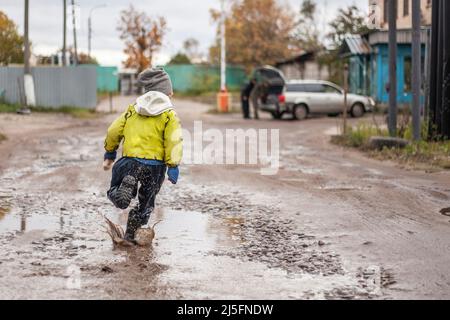 Un enfant joyeux est sale vêtements. Un petit garçon en mouvement saute dans les flaques. Un enfant méchant en automne se promène dans la boue. Bottes en caoutchouc pour sécher Banque D'Images