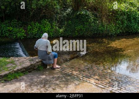 la grand-mère est assise et tient la main de son petit-fils pendant qu'il se pagaie dans un ruisseau peu profond, au soleil et à l'ombre Banque D'Images