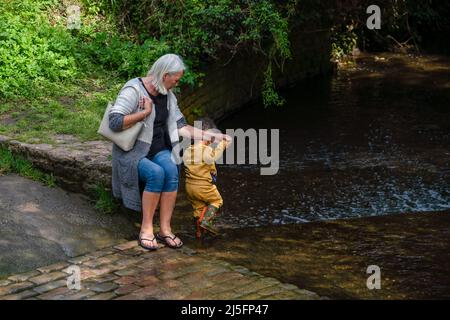 la grand-mère est assise et tient la main de son petit-fils pendant qu'il se pagaie dans un ruisseau peu profond, au soleil et à l'ombre Banque D'Images