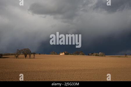 Ciel orageux spectaculaire au-dessus d'un champ labouré ensoleillé avec une ferme au loin. Banque D'Images