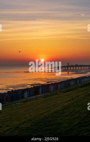 HERNE BAY : le soleil du matin qui se lève sur la mer le dimanche de Pâques avec des cabanes de plage vues en premier plan et la jetée en arrière-plan. Banque D'Images