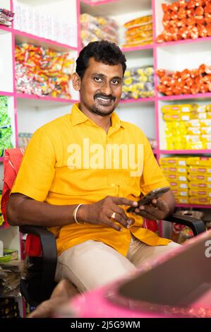 Portrait d'un homme d'affaires indien kirana ou épicerie réussi assis sur le comptoir avec le sourire en regardant la caméra - concept de petite entreprise Banque D'Images