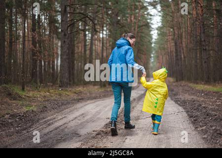 Maman et enfant marchant le long de la route de la forêt après la pluie dans les manteaux de pluie ensemble, vue arrière Banque D'Images