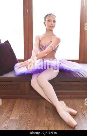 Portrait en longueur d'une belle petite ballerine dans une robe violette de performance assise près de la fenêtre et rêvant de devenir un ballet professionnel da Banque D'Images