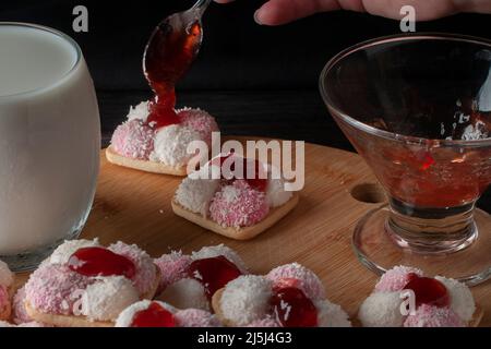 Une confiture de fraises à la main sur un guimauve et un biscuit sec de flocons de noix de coco avec un verre de lait glacé sur une planche à découper en bois Banque D'Images