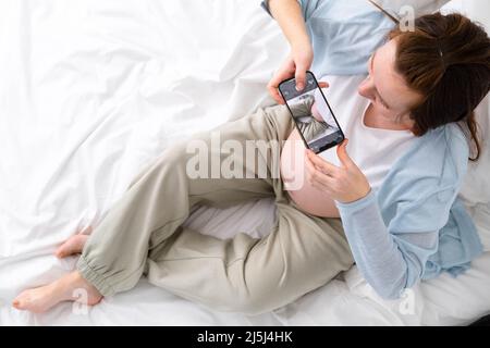 Femme enceinte expectative faisant la photo de son grand ventre au téléphone. Femme photographiant l'enfant en attente, assis sur le lit. Le grand ventre a avancé la grossesse Banque D'Images