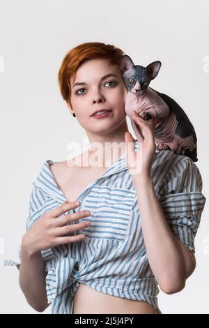 Chaton de Sphynx assis sur l'épaule d'une belle jeune femme à tête rouge de 25 ans. Portrait de femme aux cheveux courts vêtu d'une chemise à rayures blanc-bleu Banque D'Images