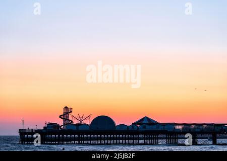 La tête de la jetée de Herne Bay Pier dans le Kent, en Angleterre, a fait une silhouette contre le ciel coloré de l'aube. Le ciel passe du violet au rouge, de l'orange au jaune et enfin au bleu plus haut. Le squelette de Helter glisse sur la jetée avec d'autres bâtiments. Banque D'Images