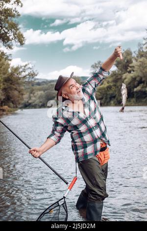 Pêcheur utilisant la pêche à la perche dans la rivière de montagne. Pêche de pêcheur sur un rever. Bon vieux pêcheur attrapé un poisson de truite. Portrait de sourire joyeux Banque D'Images