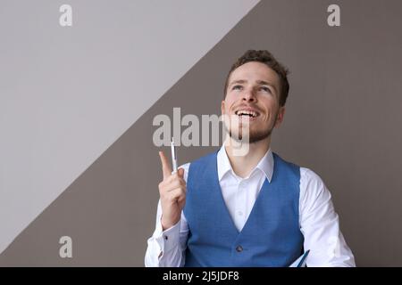 Un jeune homme d'affaires à barbe heureux se réjouit de la réussite de la pensée une idée, tient le stylet avec des expressions émotionnelles faciales sur le fond d'un mur dans le bureau. Banque D'Images