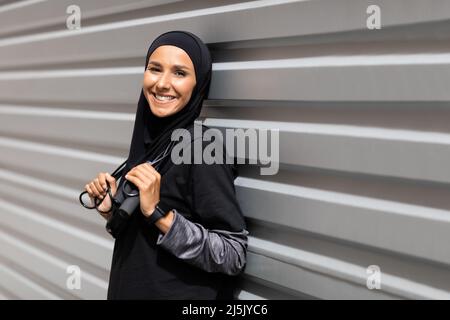 Jeune athlète souriant du Moyen-Orient dans le hijab avec corde à sauter près de fond de mur gris, espace libre Banque D'Images