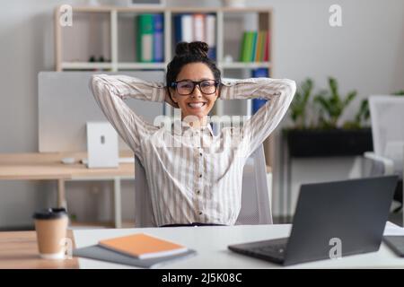 Une jeune femme heureuse qui se repose sur sa chaise, devant son ordinateur portable au bureau, se détend après une affaire réussie Banque D'Images