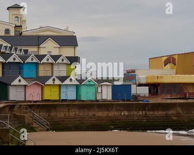 Des cabanes de plage colorées sur une colline qui se courbe à une foire d'amusement délabrée, s'enroulant autour d'une plage et d'un mur de mer parsemé de mauvaises herbes. Banque D'Images