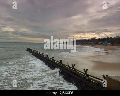 Une plage, des défenses marines, des huttes de plage et des collines à Frinton, debout contre les vagues de rupture sous un ciel orageux avec une déchirure admettant la lumière du soleil. Banque D'Images