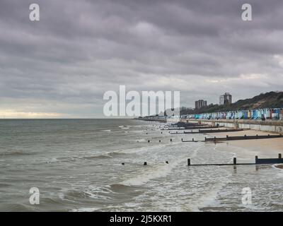 Une plage, des défenses marines, des huttes de plage et des collines à Frinton, debout contre les vagues de rupture sous un ciel orageux avec une déchirure admettant la lumière du soleil. Banque D'Images