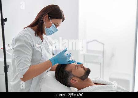 Femme dans le masque fait l'injection de remplissage pour le soulèvement des sourcils à l'homme d'âge moyen dans le salon Banque D'Images