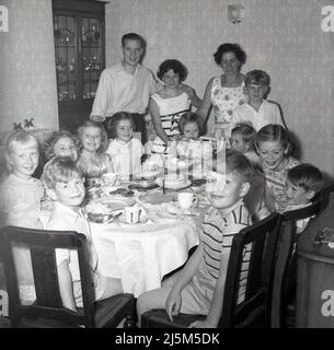 1959, historique, tous les sourires que les jeunes enfants ont le plaisir assis ensemble à une table pour une fête d'anniversaire, avec trois adultes en présence tourner leurs têtes pour une photo de groupe, Stockport, Manchester, Angleterre, Royaume-Uni. Banque D'Images