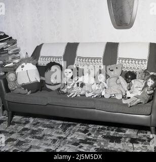 1960s, historique, une collection de jouets mous pour enfants, ours en peluche, poupées et ragdolls assis sur un canapé, Angleterre, Royaume-Uni. Le canapé a des couvertures de linge au-dessus de l'arrière, typique de cette époque, Banque D'Images