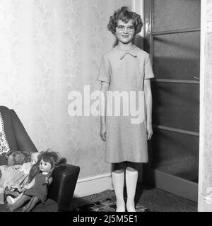 1960s, historique, une jeune fille dans une jolie robe de l'époque debout pour sa photo, ses poupées sur le canapé, Angleterre, Royaume-Uni. Banque D'Images