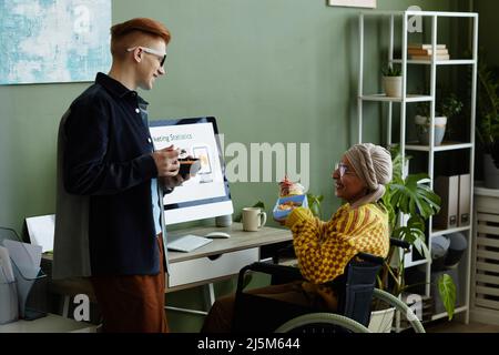 Portrait de la jeune femme en fauteuil roulant au bureau et en train de discuter avec son collègue pendant le déjeuner Banque D'Images