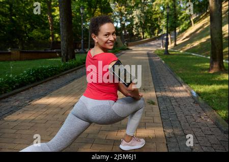 Vue du dos à une femme africaine en train de faire des fentes tout en faisant de l'exercice en plein air, sourit avec un sourire éclatant en regardant l'appareil photo, en appréciant lui Banque D'Images